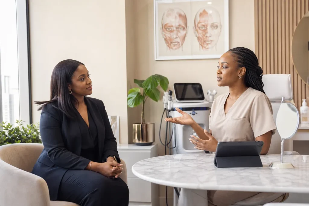 A Kenyan woman and a clinician have a detailed discussion in a private, modern consultation room equipped with anatomical charts and aesthetic equipment.