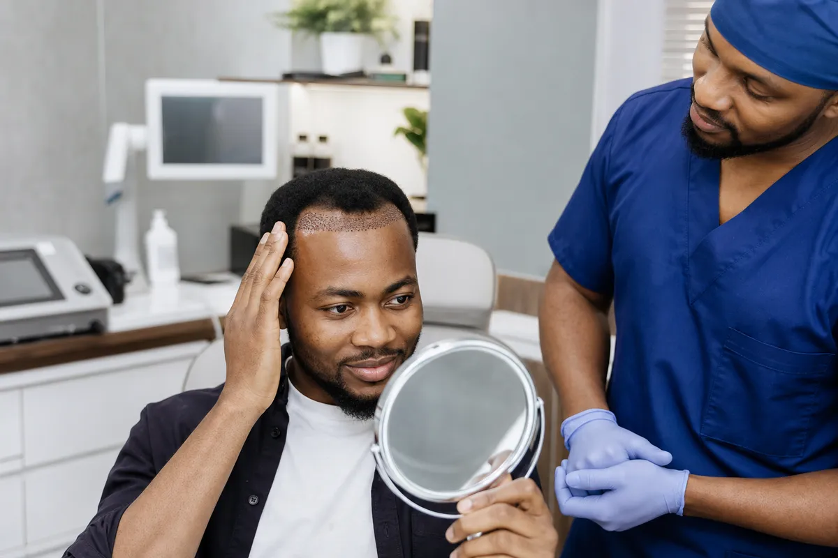 A Kenyan man with a newly restored hairline examines his reflection in a mirror as a supportive medical professional stands nearby in a modern clinic.