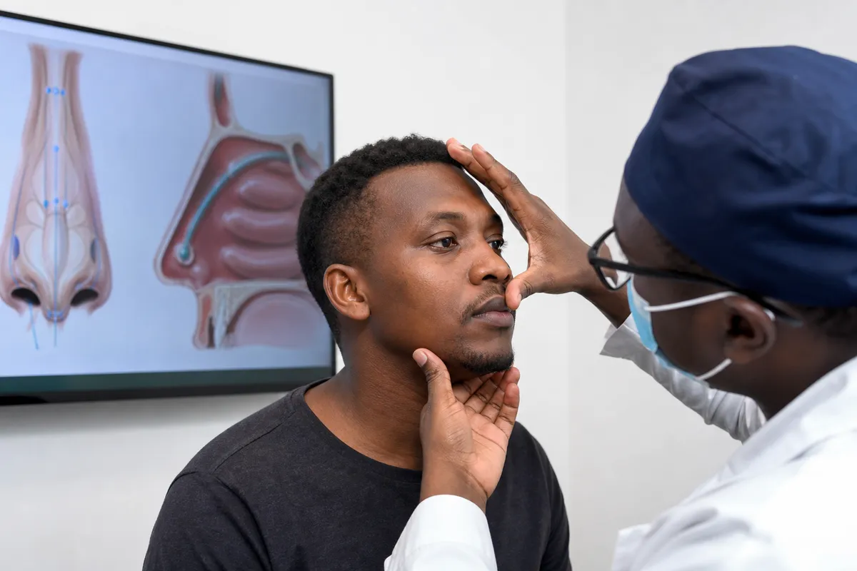A surgeon gently evaluates the nose of a Kenyan man while referencing nasal diagrams on a screen in a clinical setting.