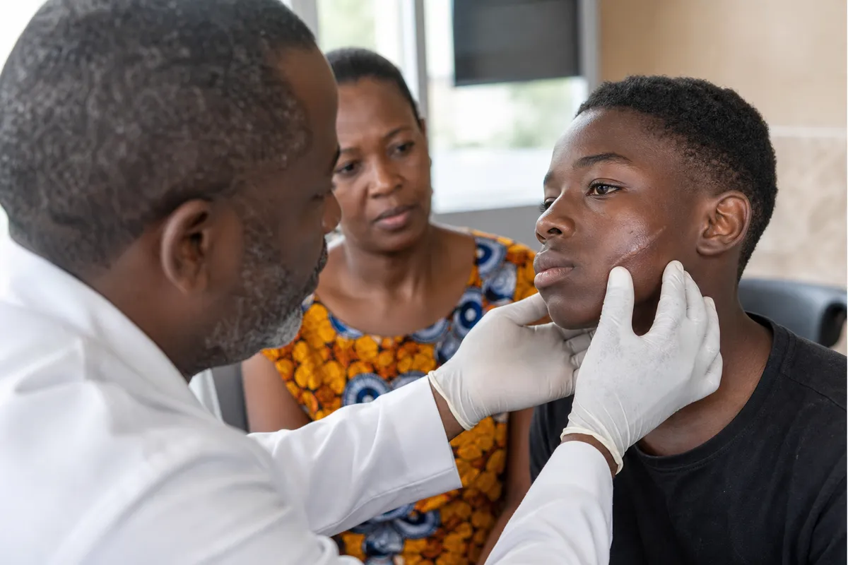 A Kenyan plastic surgeon examines a teenage boy’s healed facial scar while a parent watches supportively in a modern clinic setting.