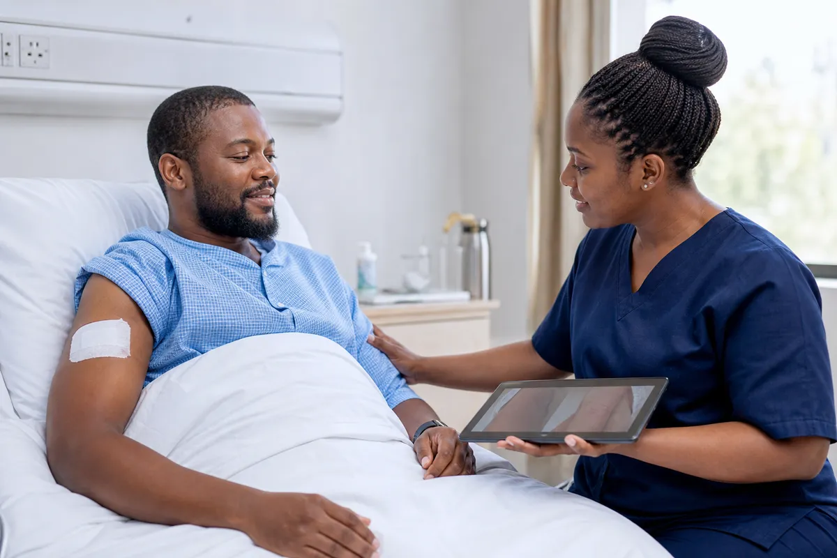 A Kenyan man reclines in a private clinic room while a nurse checks a small dressing on his upper arm and discusses wound aftercare using a tablet.