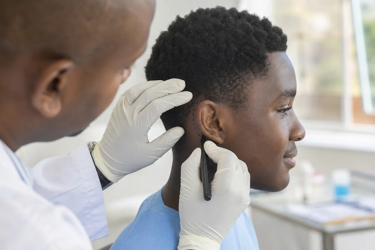 A Kenyan plastic surgeon with gloved hands carefully marks the ear of a teenage boy for otoplasty preparation in a well-lit clinic, while the teen appears calm and attentive.