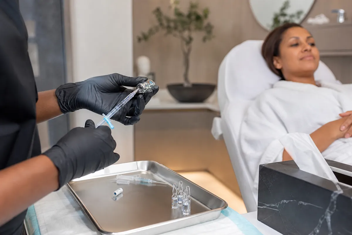 Gloved hands prepare hyaluronic acid dermal filler syringes with vials on a metal tray in a clean treatment room, as a Kenyan woman reclines in the background.