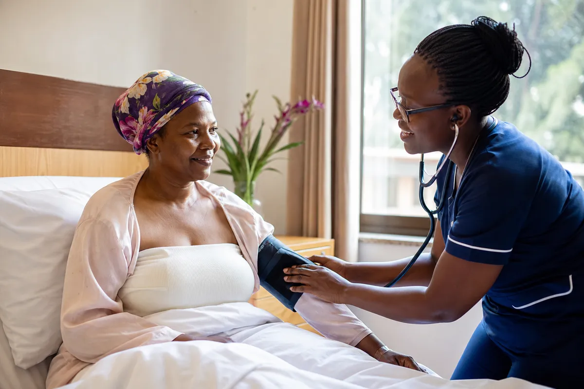 A Kenyan woman in a clinic bed gently smiles as a nurse checks her vitals in a softly lit, comfortable recovery room.